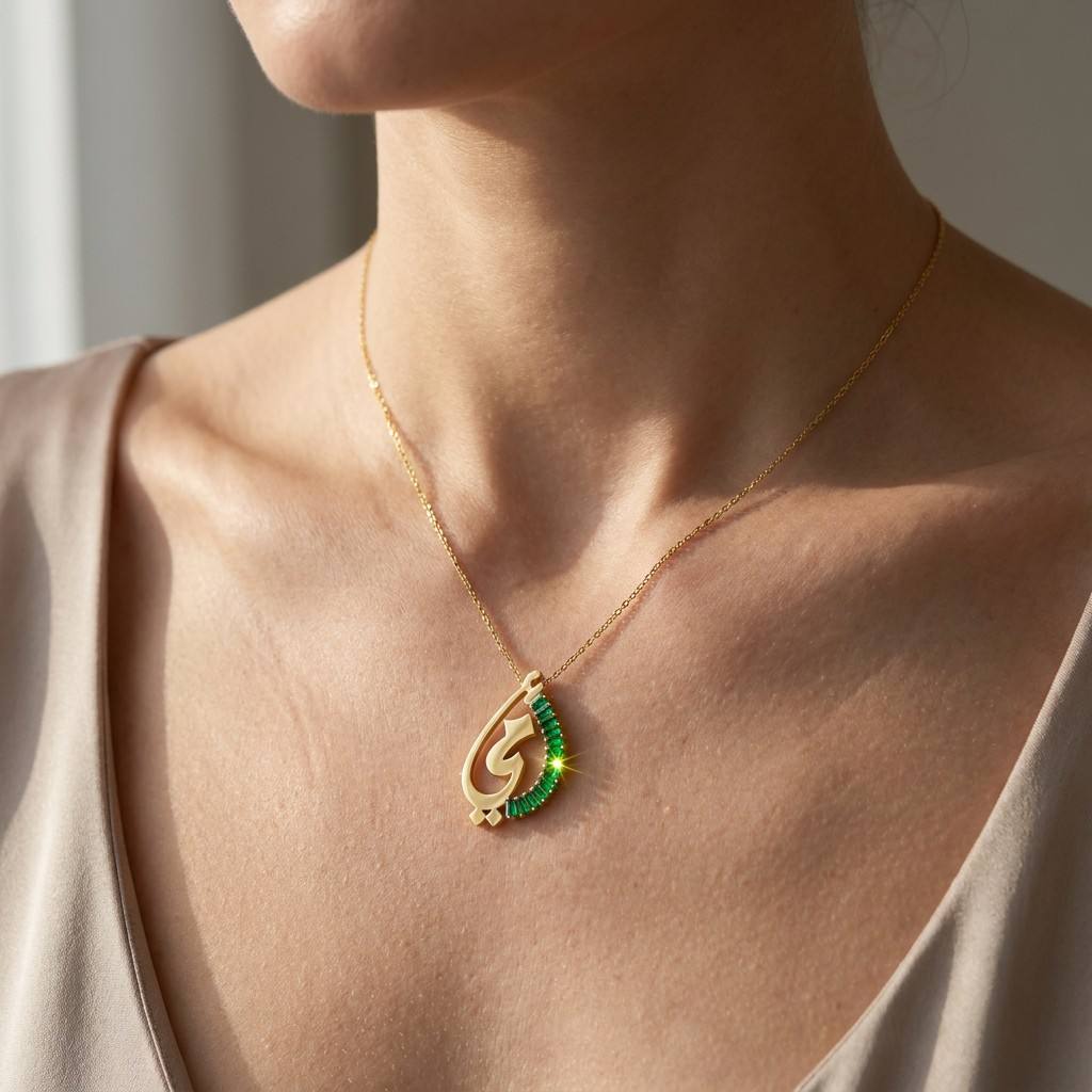 Model wearing gold Arabic letter pendant necklace with green gemstone accent on neckline