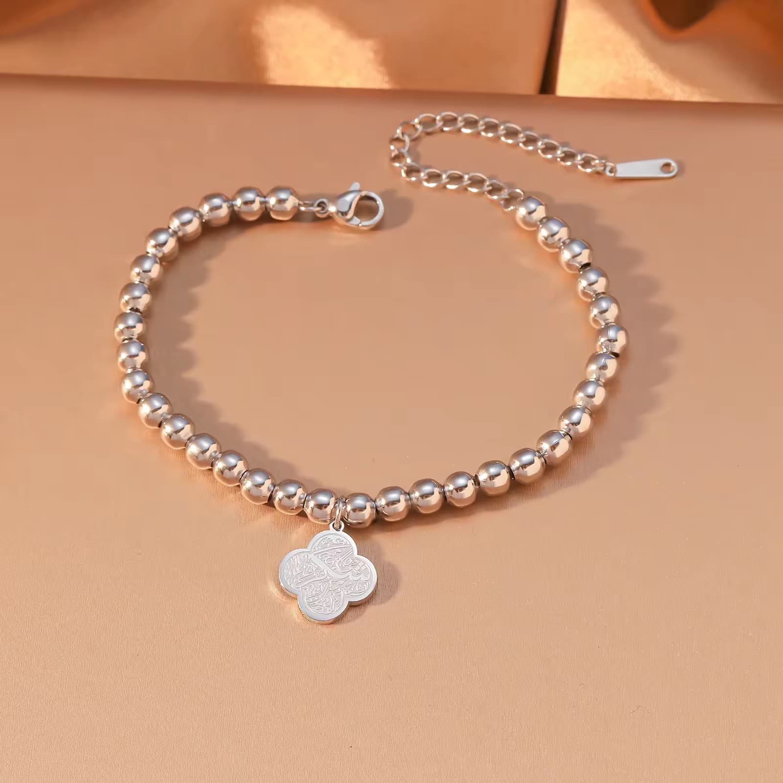 CIGAONI Clover of Virtue polished silver beaded bracelet flat lay, quatrefoil charm with Arabic calligraphy engraving, lobster clasp and extender chain on beige background