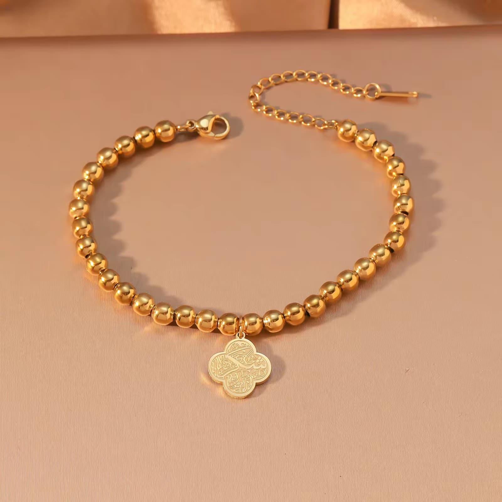 CIGAONI Clover of Virtue 18K gold beaded bracelet flat lay, quatrefoil charm with Arabic calligraphy engraving, lobster clasp and extender chain on beige background