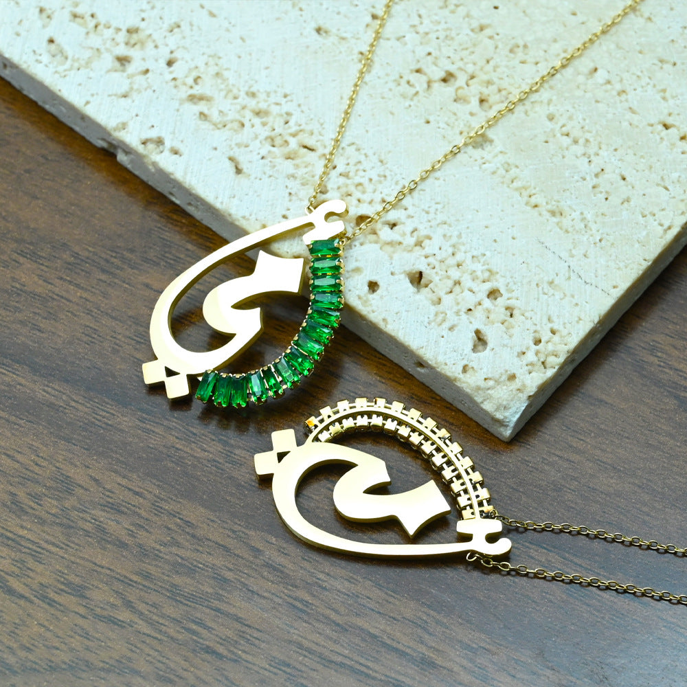 Close-up of gold Arabic script charm necklace with emerald green crystal detail on teal background